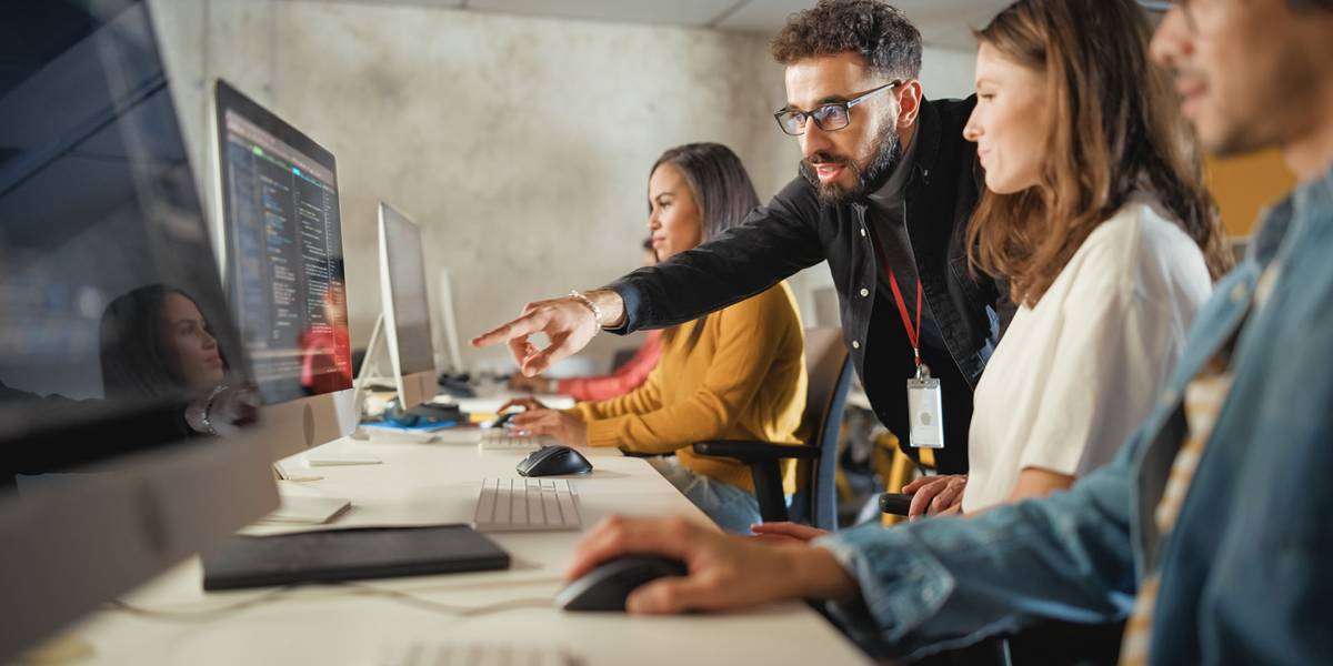 Führungskraft gibt einem Teammitglied Anweisungen am Computerarbeitsplatz, während weitere Personen mitarbeiten.