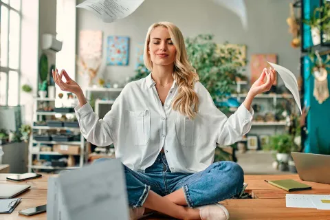 Resiliente Mitarbeiterin meditiert auf ihrem Bürotisch. Um sie herum wehen verschiedene Unterlagen. © Adobe Stock, Vasyl