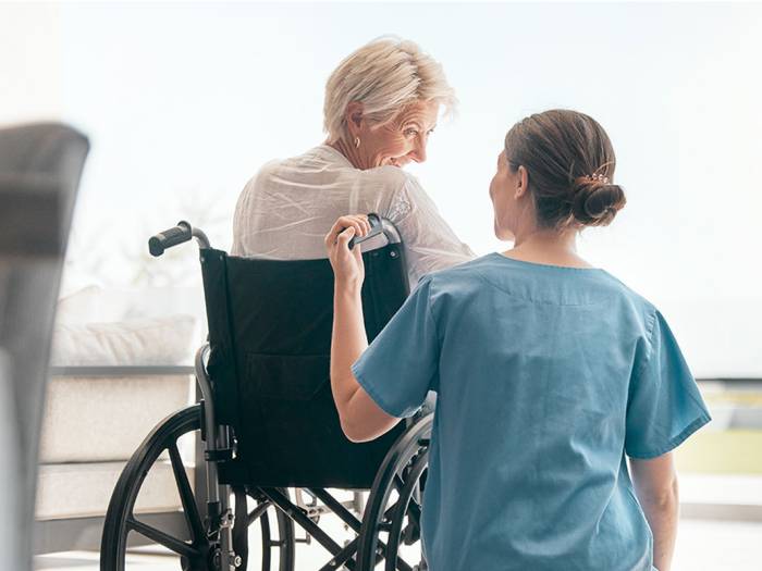 Pflegekraft schiebt Seniorin im Rollstuhl und spricht mit ihr in einem hellen Pflegezimmer.