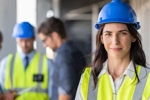 Drei Männer besprechen im Hintergrund Schutzmaßnahmen nach dem STOP-Prinzip und Frau schaut im Vordergrund selbstsicher mit persönlicher Schutzausrüstung in Richtung des Betrachters. © Adobe Stock, Rido