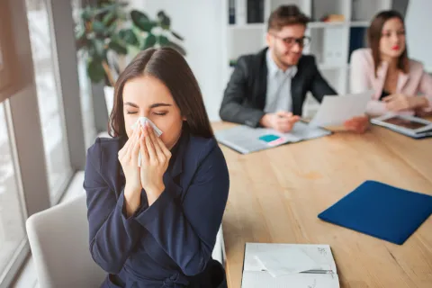Mitarbeiterin mit Grippe dreht sich von ihren Kollegen weg und schnaubt die Nase. Eine Impfung kann sie schützen. © Shutterstock, Estrada Anton