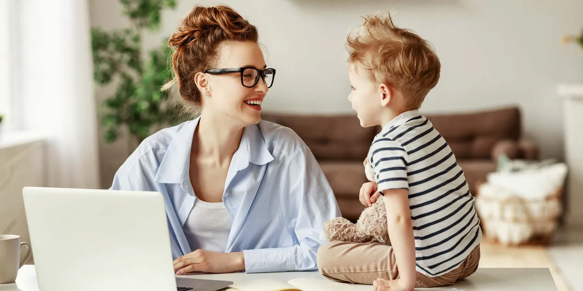 Eine Mutter vereinbart Beruf und Familie und lächelt beim Arbeiten ihr Kind an. © Adobe Stock, JenkoAtaman