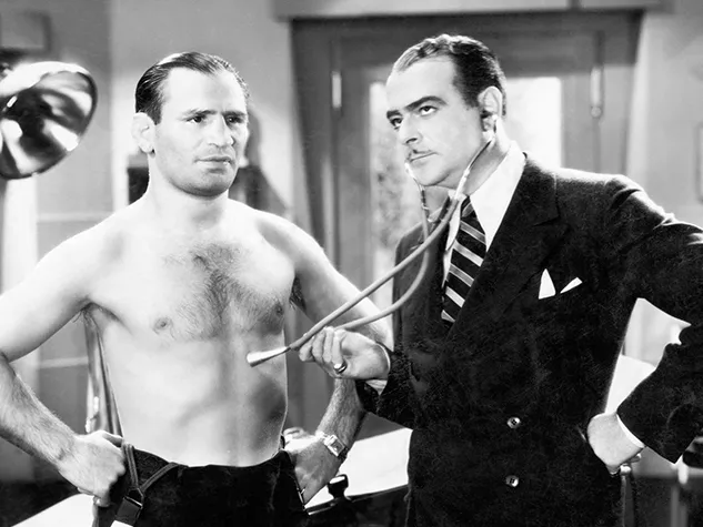 Schwarzweißbild eines Mannes, der vom Arzt untersucht wird. Heute sind Betriebsärzte eine Selbstverständlichkeit, das war allerdings nicht immer so. © Shutterstock, Everett Collection