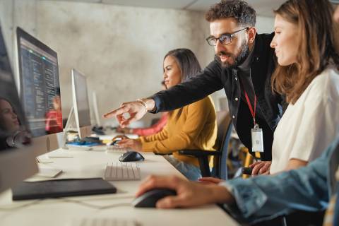 Führungskraft gibt einem Teammitglied Anweisungen am Computerarbeitsplatz, während weitere Personen mitarbeiten.