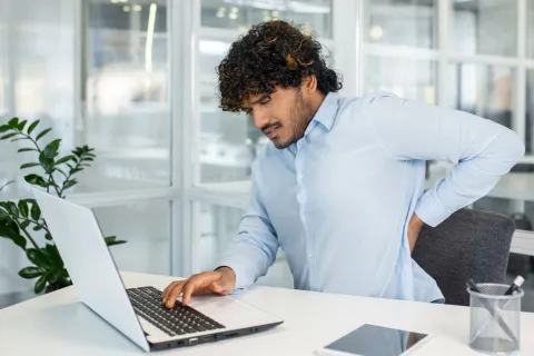 Ein junger Berufstätiger leidet unter Rückenschmerzen, während er in einem modernen Büro an seinem Laptop arbeitet. Das Bild fasst häufige Gesundheitsprobleme am Arbeitsplatz zusammen. © Adobe Stock, Liubomir