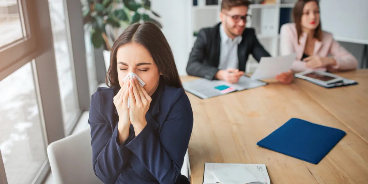Mitarbeiterin mit Grippe dreht sich von ihren Kollegen weg und schnaubt die Nase. Eine Impfung kann sie schützen. © Shutterstock, Estrada Anton
